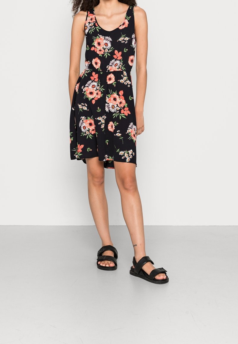 Femme portant une robe noire sans manches avec un imprimé floral rose et blanc et des sandales noires ouvertes à l'avant, debout devant un mur clair uni.