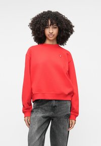 Κόκκινη μπλούζα με στρογγυλή λαιμόκοψη, χαμηλωμένους ώμους και ριμπ μανσέτες. Διαθέτει ένα μικρό λογότυπο στο στήθος. Φοριέται με ξεθωριασμένο γκρίζο τζιν.