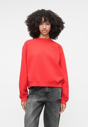 Rød sweatshirt med rund halsudskæring, sænkede skuldre og ribstrikkede manchetter. Funktionen inkluderer et lille logo på brystet. Båret med slidte grå jeans.