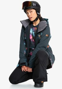 Nainen, joka on pukeutunut mustaan Roxy hiihtokypärään, värikkääseen tie-dye-paitaan, tummaan teal-takkiin ja mustiin lumihousuihin, polvistuu kädet yhteen puristettuna.