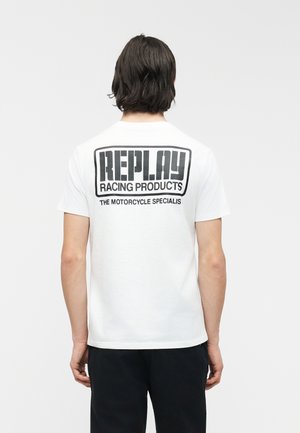 Persoon met donker haar van gemiddelde lengte staat met de rug naar de camera, draagt een wit T-shirt met zwarte tekst "REPLAY RACING PRODUCTS THE MOTORCYCLE SPECIALIS".