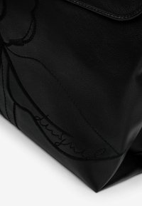 Musta nahkalaukku teksturoidulla pinnalla, jossa on kirjailtu kukkakuvio ja logo. Jäykkä muoto, läppäkiinnitys. Ei näkyvää metalliosaa.