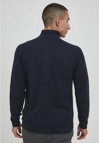 Marineblauer strukturierter Pullover mit hohem Kragen, ausgestattet mit einem gerippten Saum und Bündchen. Strickmuster und figurbetontes Design von hinten sichtbar.