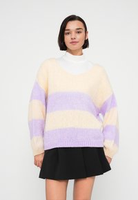 Pull à rayures pastel dans des tons crème et lavande, avec une coupe ample, un col en V et un ourlet côtelé. Porté sur un col roulé blanc.