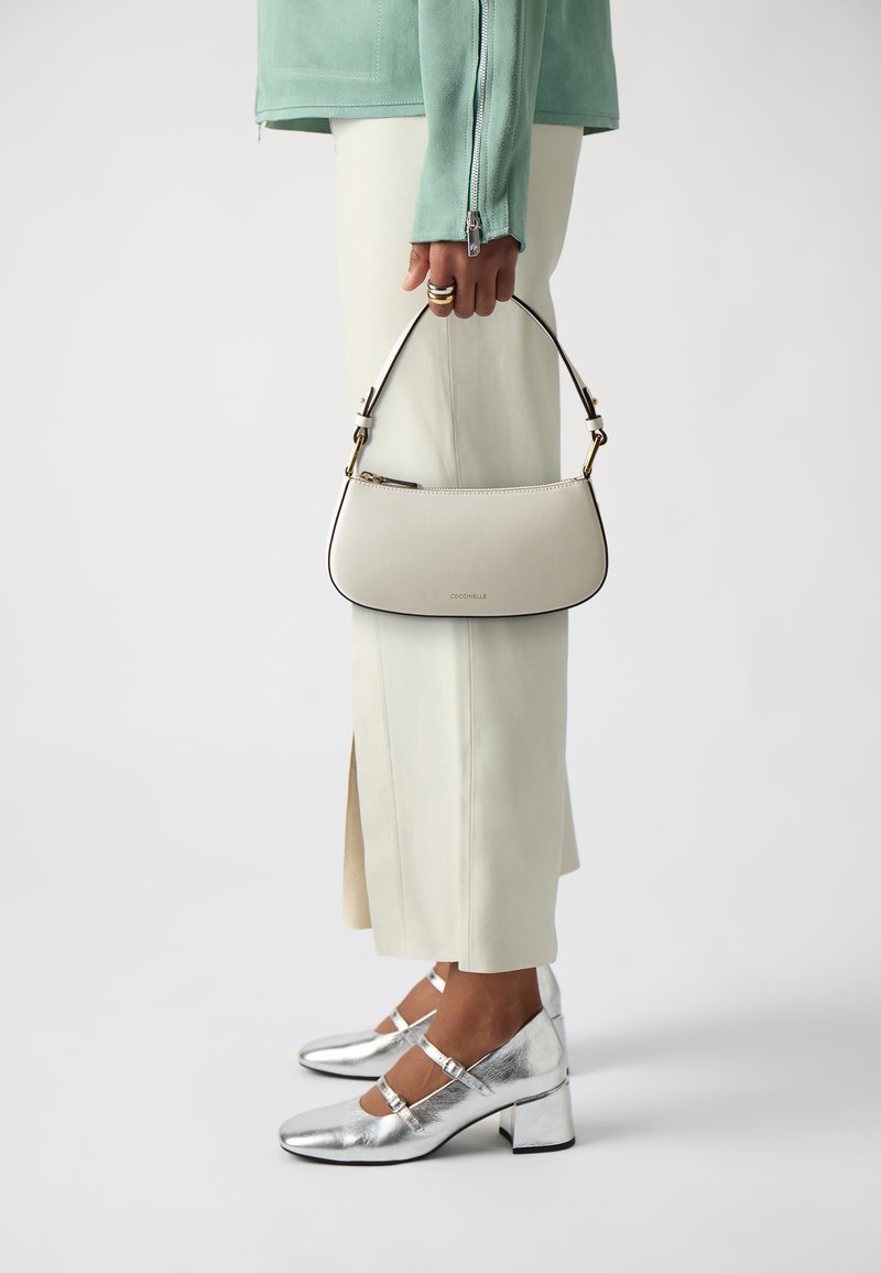 Piccola borsa strutturata in pelle beige chiaro con rifiniture nere, con hardware dorato. Il modello indossa tacchi metallici argento e pantaloni cream sartoriali.