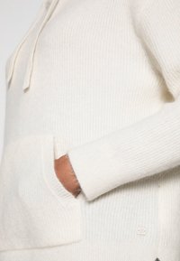 Hoodie lavorato a maglia di colore crema con superficie testurizzata, tasca frontale e cordino piatto. Presenta un piccolo etichetta di marca nella parte inferiore.