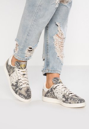 Persoon draagt lichtblauwe gescheurde spijkerbroek en grijs-witte sneakers met slangenhuidpatroon, witte zolen en veters.