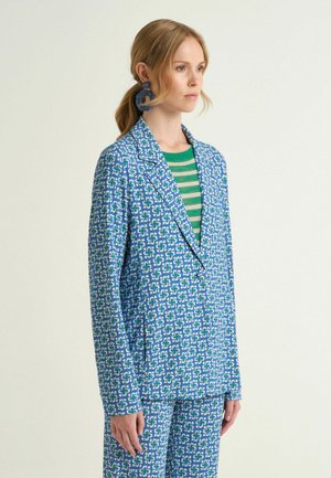 Donna con capelli biondi raccolti in una coda bassa, indossa un blazer con motivo blu e pantaloni abbinati su un maglione a righe verdi e grigie, in piedi di profilo.