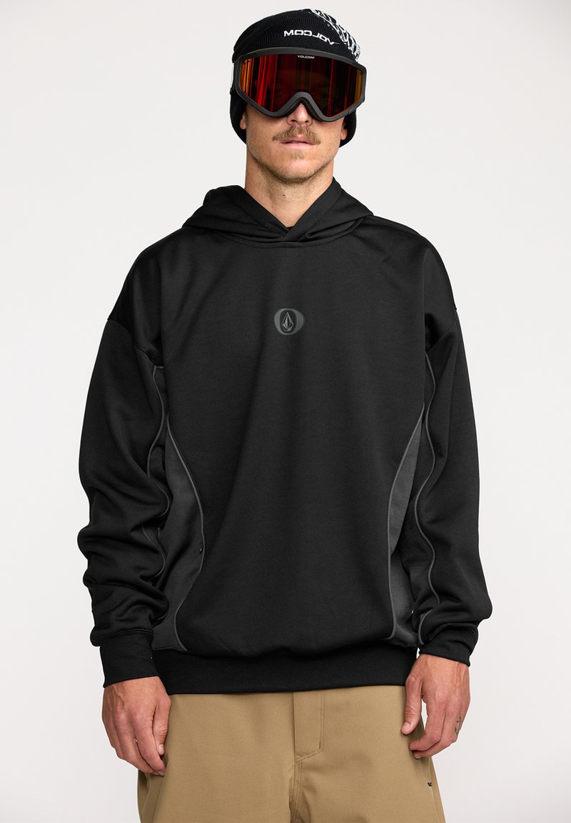 Hoodie noir avec un logo sur le devant, présentant une coupe décontractée, un tissu texturé et des panneaux latéraux gris contrastants. Associé à des lunettes de ski et un bonnet.