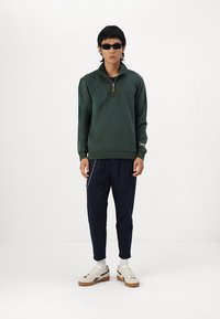 Suéter verde con cremallera en el cuello y puños acanalados, combinado con pantalones ajustados azul marino que presentan un detalle de cadena. El calzado son zapatillas blancas con suelas marrones.