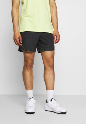 Shorts en denim noir avec un ourlet effiloché, portés avec un t-shirt léger vert clair. Des baskets blanches et des chaussettes blanches complètent la tenue.