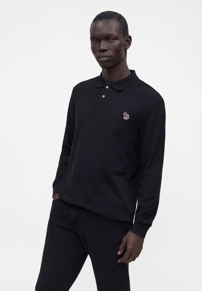 Homme portant un polo noir à manches longues avec un petit logo zèbre coloré sur la poitrine, debout avec une main dans la poche.