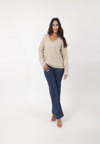 Beige gestrickter Pullover mit V-Ausschnitt und Fronttasche, kombiniert mit dunklen, ausgestellten Jeans. Das Model trägt hellbeige Riemchen-Absatzschuhe.
