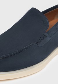 Zapato de charol azul marino sin cordones con acabado de ante texturizado, suela de goma beige, costuras decorativas en la parte superior y diseño sin costuras.