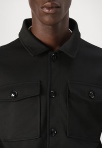 Camisa preta de botão com uma superfície texturada, dois bolsos frontais e colarinho. Apresenta botões pretos e um design limpo e estruturado.