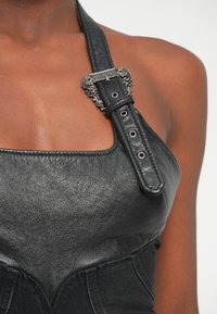 Close-up de uma pessoa a usar um top de couro preto com um fecho decorativo prateado a prender uma alça larga sobre o ombro.