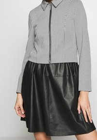 Veste à motifs noir et blanc avec un col à revers et une fermeture éclair, associée à une jupe en cuir noir unie. Texture lisse, haut ajusté.