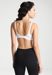 Hvit bh med nettingdetaljer, justerbare stropper og hækle-lukking. Modellen har på seg svarte høytlivs leggings.