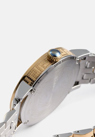 Orologio in metallo con finitura oro e argento, lunetta in oro texturizzato, corona con gemma blu e bracciale in metallo a maglie. Il retro presenta un design inciso.