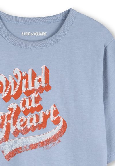 T-shirt bleu clair avec texte vintage rouge et blanc "Wild at Heart" et petit logo Zadig & Voltaire à l'intérieur du col et sous le texte.