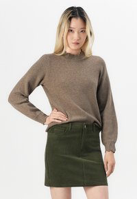 Brun strikkegenser med volangkant på halsen og mansjettene, parret med et grønt cordfløyels mini-skjørt med frontlommer og knappelukking.