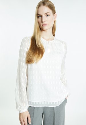 Junge Frau mit langen blonden Haaren, die eine weiße, strukturierte Bluse und graue Hosen trägt, steht mit einer Hand in der Tasche.
