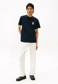 Camiseta de algodón azul marino con cuello redondo, que presenta un pequeño logo bordado. Combinada con pantalones ajustados blancos y zapatos negros.
