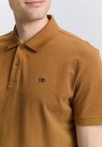 Brun polo-skjorte lavet af tekstureret stof med krave, der har en to-knap lukning og et lille, mørkt logo på brystet.