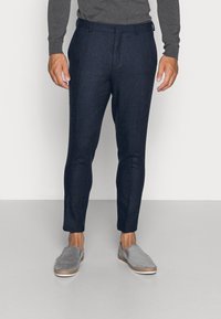 Muž ve tmavě modrých kalhotách na míru, šedém dlouhém topu a šedých slip-on botách, stojící na světle šedé podlaze před bílou zdí.