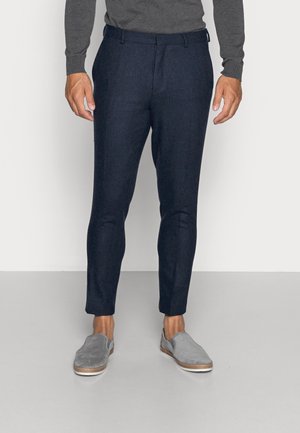 Mann in navyblauen maßgeschneiderten Hosen, grauem Langarmoberteil und grauen Slippern, der auf einem hellgrauen Boden vor einer weißen Wand steht.