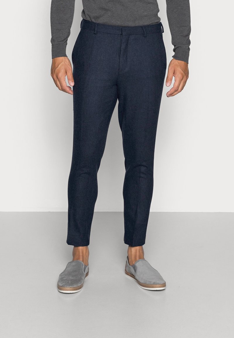 Muž ve tmavě modrých kalhotách na míru, šedém dlouhém topu a šedých slip-on botách, stojící na světle šedé podlaze před bílou zdí.