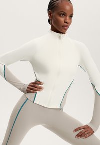 Jachetă albă cu fermoar, cu accente tei, asociată cu colanți bej ajustați, ambele având o textură elegantă și netedă, precum și un design athletic.