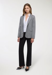 Blazer grigio con texture morbida, rever a lancia e tasca frontale, abbinato a una camicetta bianca e pantaloni neri, completato da décolleté a punta.