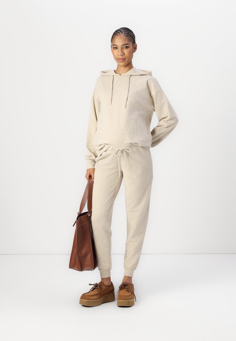 Beige Kapuzenpullover mit Kordelzug, passende Jogginghose und braune Plateauschuhe. Braune Handtasche mit Schultergurt. Glatte Stofftextur.
