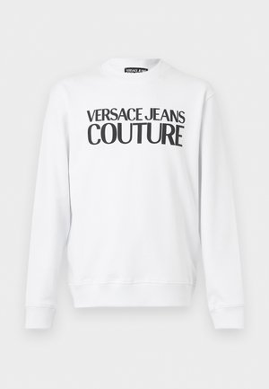 Bijela majica dugih rukava s okruglim izrezom i crnim natpisom "Versace Jeans Couture" preko prsa.