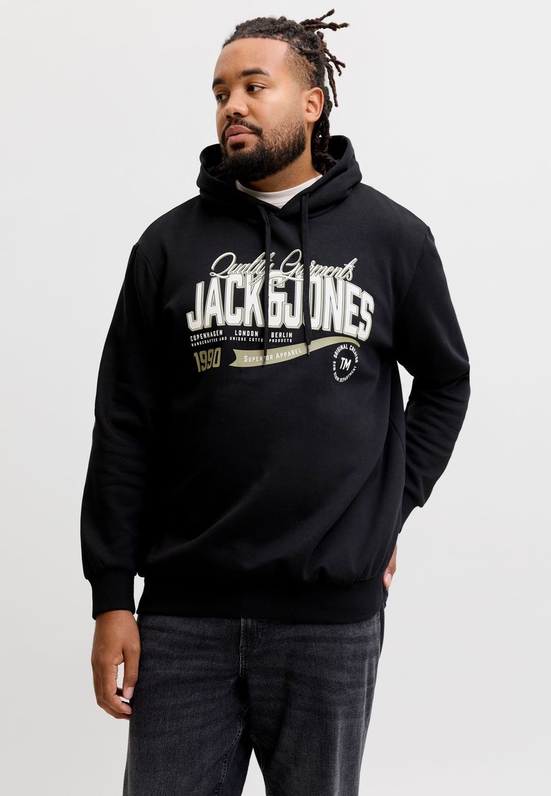 Homme aux cheveux tressés portant un sweat à capuche noir Jack & Jones et un jean foncé, regardant sur le côté avec une main dans la poche.