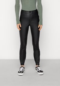 Calças pretas em pele sintética com um corte slim e textura suave, combinadas com uma blusa verde de manga longa com nervuras e ténis pretos.