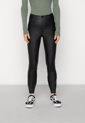 Calças pretas em pele sintética com um corte slim e textura suave, combinadas com uma blusa verde de manga longa com nervuras e ténis pretos.
