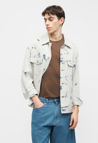 Νέος άνδρας με κοντά καστανά μαλλιά, που φοράει ένα λευκό και μπλε τζιν μπουφάν με σχέδιο tie-dye, καφέ πουκάμισο και μπλε τζιν, κοιτάζει αριστερά με το ένα χέρι στην τσέπη.