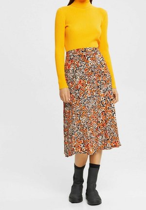 Femme portant un haut jaune à manches longues côtelé, une jupe midi fleurie avec des fleurs orange, et des bottines noires, debout devant un fond uni.