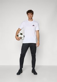 Hvid atletisk t-shirt med sorte og rosa detaljer, parret med sorte træningsbukser med hvide sidelister. Holder en sort og hvid fodbold.