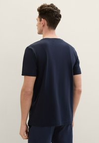 Hombre con cabello rizado corto que lleva una camiseta de manga corta azul marino lisas y pantalones a juego, de pie de espaldas en un fondo neutro.