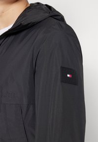 Sort hættetrøje med lynlåslukning foran, der har et Tommy Hilfiger logo-patch på den øvre ærme.