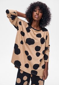 Beżowy top z długim rękawem z dużymi czarnymi grochami, okrągłym dekoltem i luźnym krojem. Materiał wydaje się miękki o gładkiej fakturze.