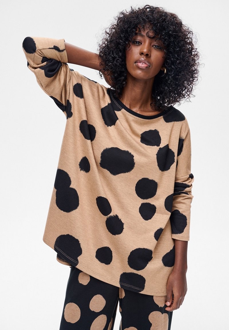 Beżowy top z długim rękawem z dużymi czarnymi grochami, okrągłym dekoltem i luźnym krojem. Materiał wydaje się miękki o gładkiej fakturze.