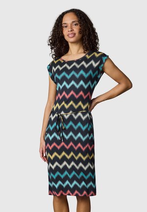 Kvinde med krøllet hår iført en knælængde sort kjole med farverige zigzag-mønstre og snøre i taljen, poserer med den ene hånd på hoften.
