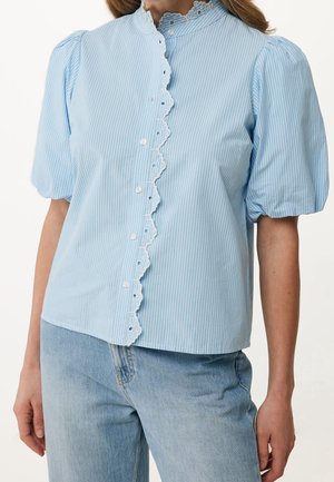 Lyseblå bluse med hvide lodrette striber, bølget blondekant langs knapstolpen, puffede korte ærmer, kombineret med lyseblå jeans.