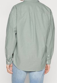 Chemise vert clair à manches longues avec un col, des poignets à boutons et un ourlet arrondi. Présente un empiècement au dos avec des plis pour un meilleur ajustement.