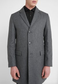 Grijze wollen jas met een getailleerde pasvorm, enkel-breasted ontwerp, notch revers, twee frontzakken en zwarte knoppen. Gekleed over een zwart shirt.