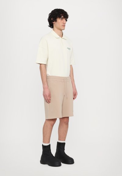 Polo shirt beige clair à manches courtes, associé à un short beige avec une taille élastique. Bottines noires à surface texturée.